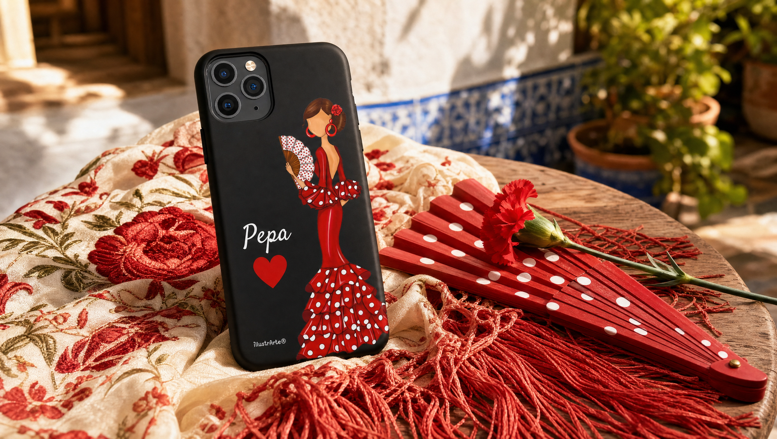 Móvil con la funda personalizada Flamenca Pepa de illustrArte, con el nombre Pepa, sobre un mantón de Manila junto a un abanico de lunares y un clavel rojo en un patio andaluz